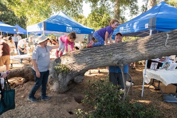 Two small children climbing a tree branch outdoors with two adults guiding them. LADWP blue canopies are in the background. 