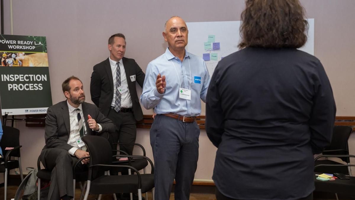 Four people talking in a room with a white board with sticky notes and event poster in the background. 