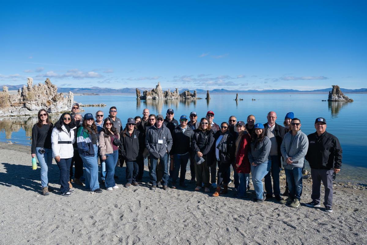 A group of people smiling and posing outdoors for a photo in front of a large body of water with rock-like formations, clouds, and mountains in the background.