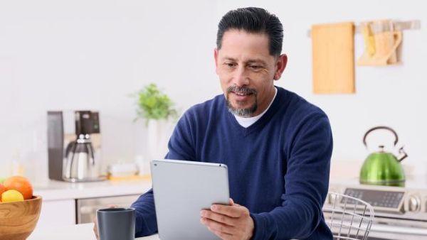 Man in blue sweater sitting in a chair holding and looking at an iPad in one hand, with his other hand on a mug.