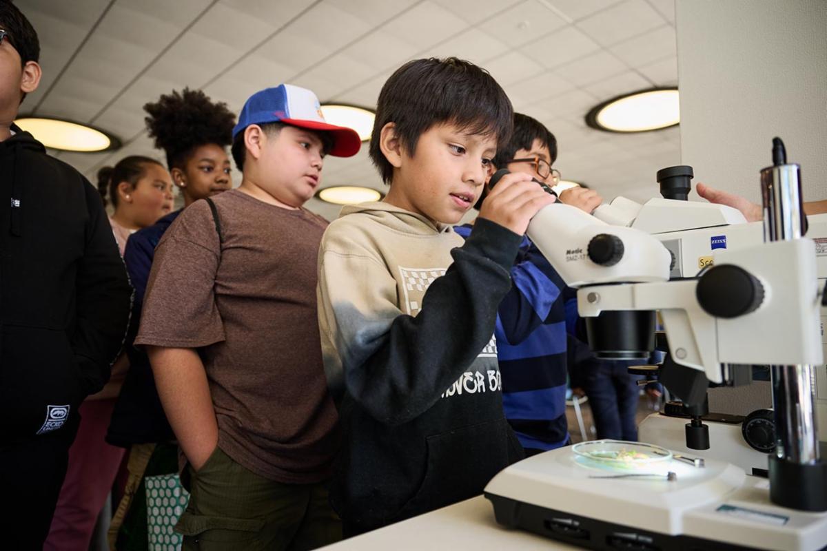 Young male student looking inside a microscope with other students lined up behind him.
