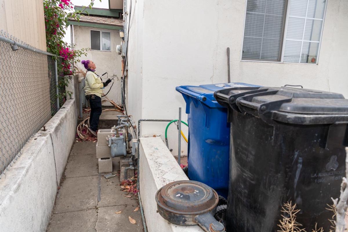 Female utility worker next to house looking at equipment with recycle and trash bins in sight.