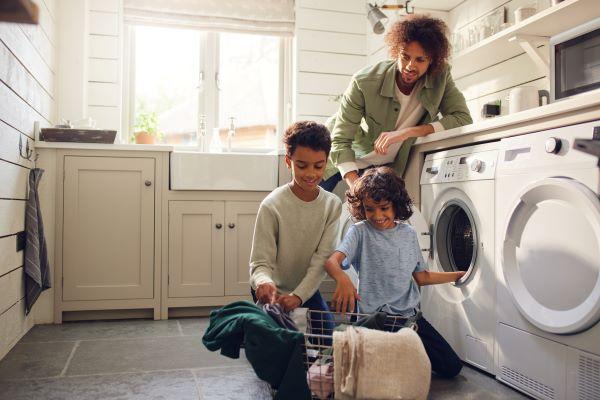Two young children taking laundry out of a dryer machine into a basket while an adult leans over the dryer machine looking at them.