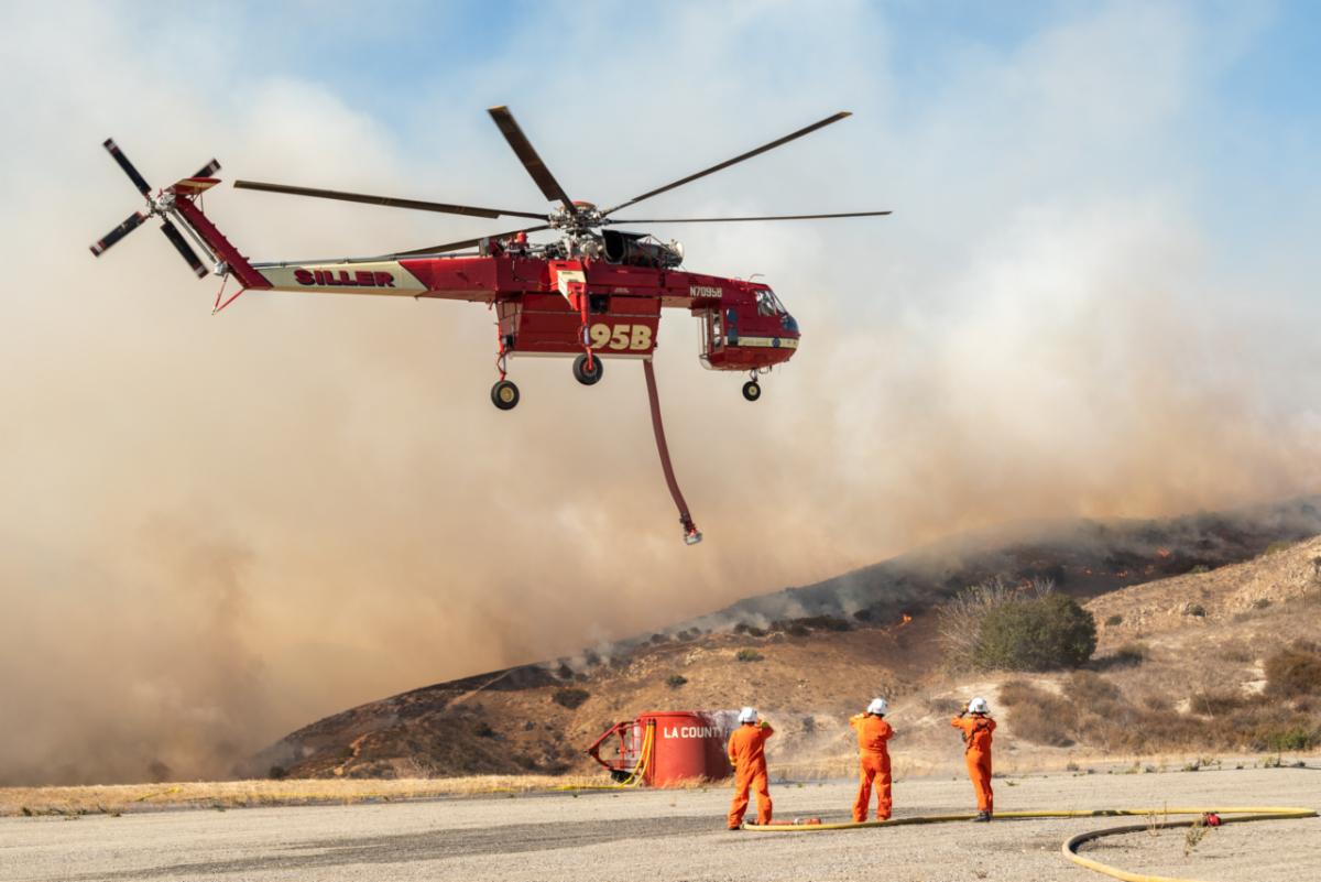 Image of a helicopter collecting water from a water tank to help put out a wildfire there are three people standing nearby on the ground as support