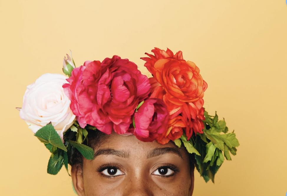 an image of a brown skinned femme-presenting person with only the top half of their face shown. a yellow background and a crown of while, red, and orange flowers around their head