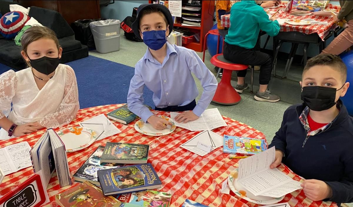 Three elementary school students sit at a table with a red and white table cloth, surrounded by books and menus for a "book tasting," as part of a Read Across America celebration.