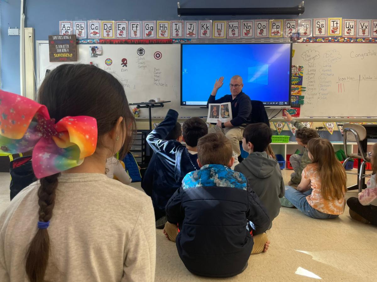 A male officer of the Middletown Township Police Department is surrounded by Lincroft Elementary School students while reading a book to the group.