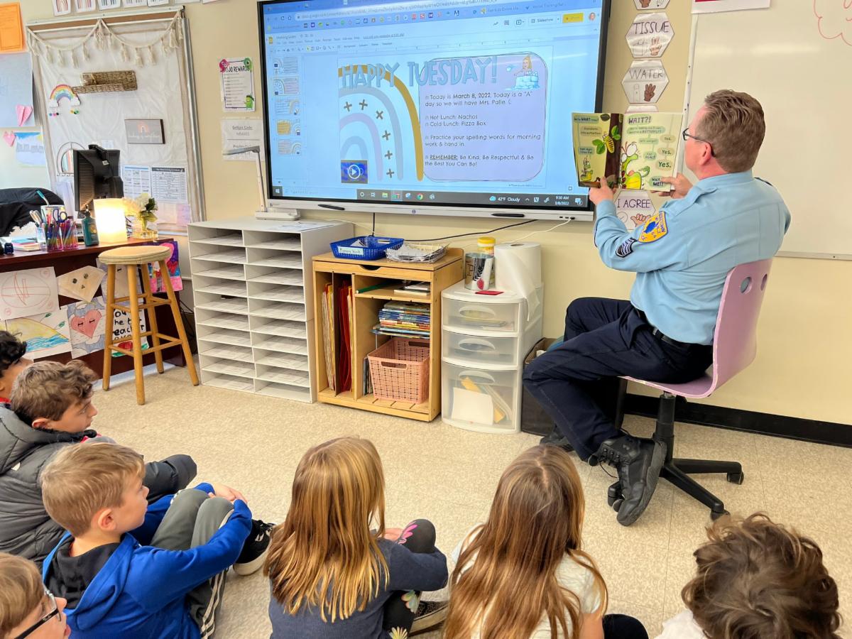A male officer of the Middletown Township Police Department is surrounded by Lincroft Elementary School students while reading a book to the group.