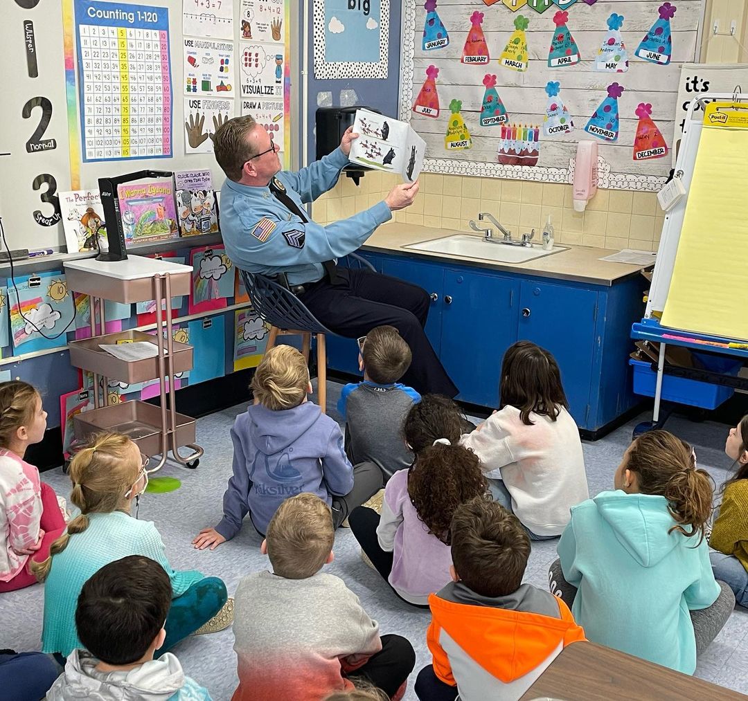 A male officer of the Middletown Township Police Department is surrounded by Lincroft Elementary School students while reading a book to the group.