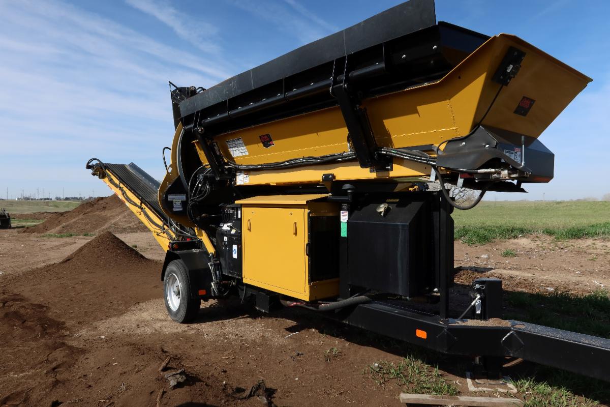 Large black and yellow trommel screener equipment, with a pile of screened compost behind
