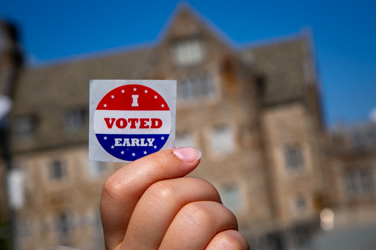A hand holding an I Voted Early sticker with a Duke building in the background