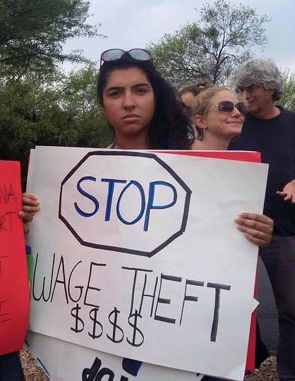 Young woman with brown skin and black hair holding a poster that says STOP Wage Theft.
