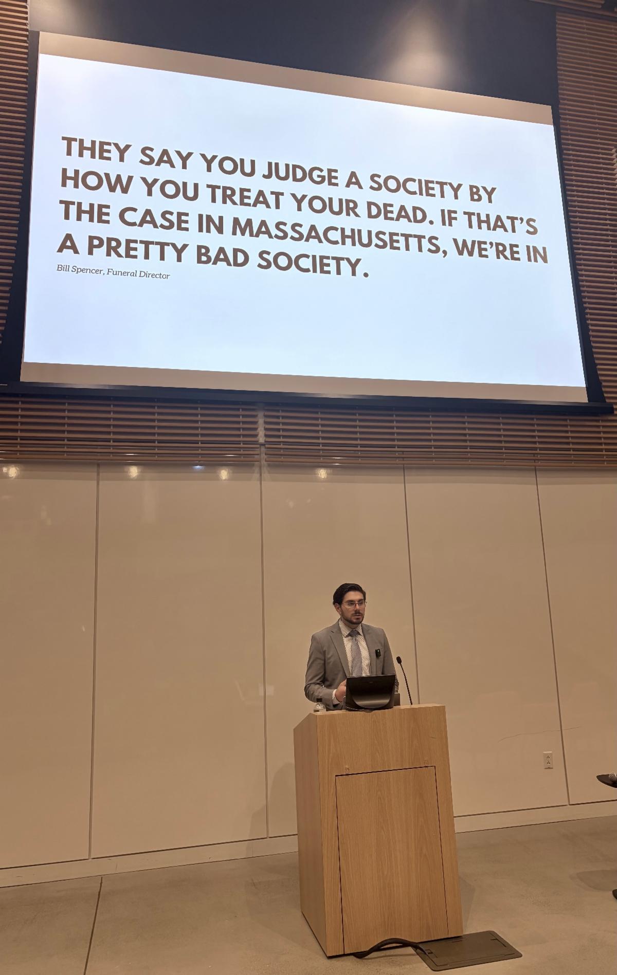 Man standing at a podium in front of a slideshow that says "They say you judge a society by how you treat your dead. If that's the case in Massachusetts, we're in a pretty bad society."