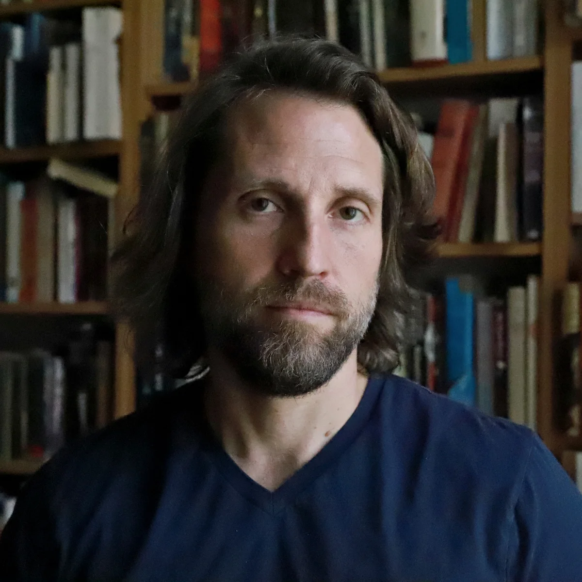 A white man with shoulder-length brown hair and facial hair. He is looking directly into the camera with a serious facial expression.