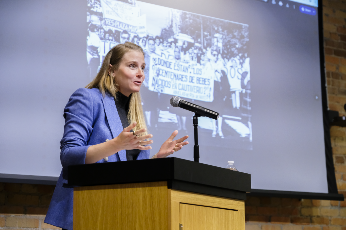 Haley Cohen Gilliland speaking at a podium with a microphone. A black and white photo of women marching with a banner is projected behind her.