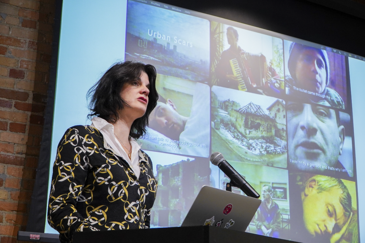 Nora Krug standing at a podium with a microphone, with her illustrations projected behind her