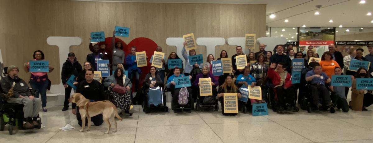 Group of 100 advocates in front of I Love NY sign. The photo shows people holding signs reading Fund Access to Home , CDPA is Essential, and Support Independent Living Funding while some chanted with fists in the air! 