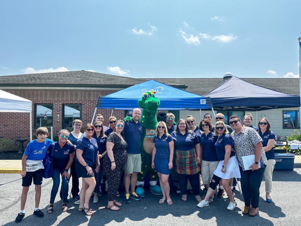 Image of Sail Staff standing under a pop up tent with a gigantic green dinosaur on a bright sunny day