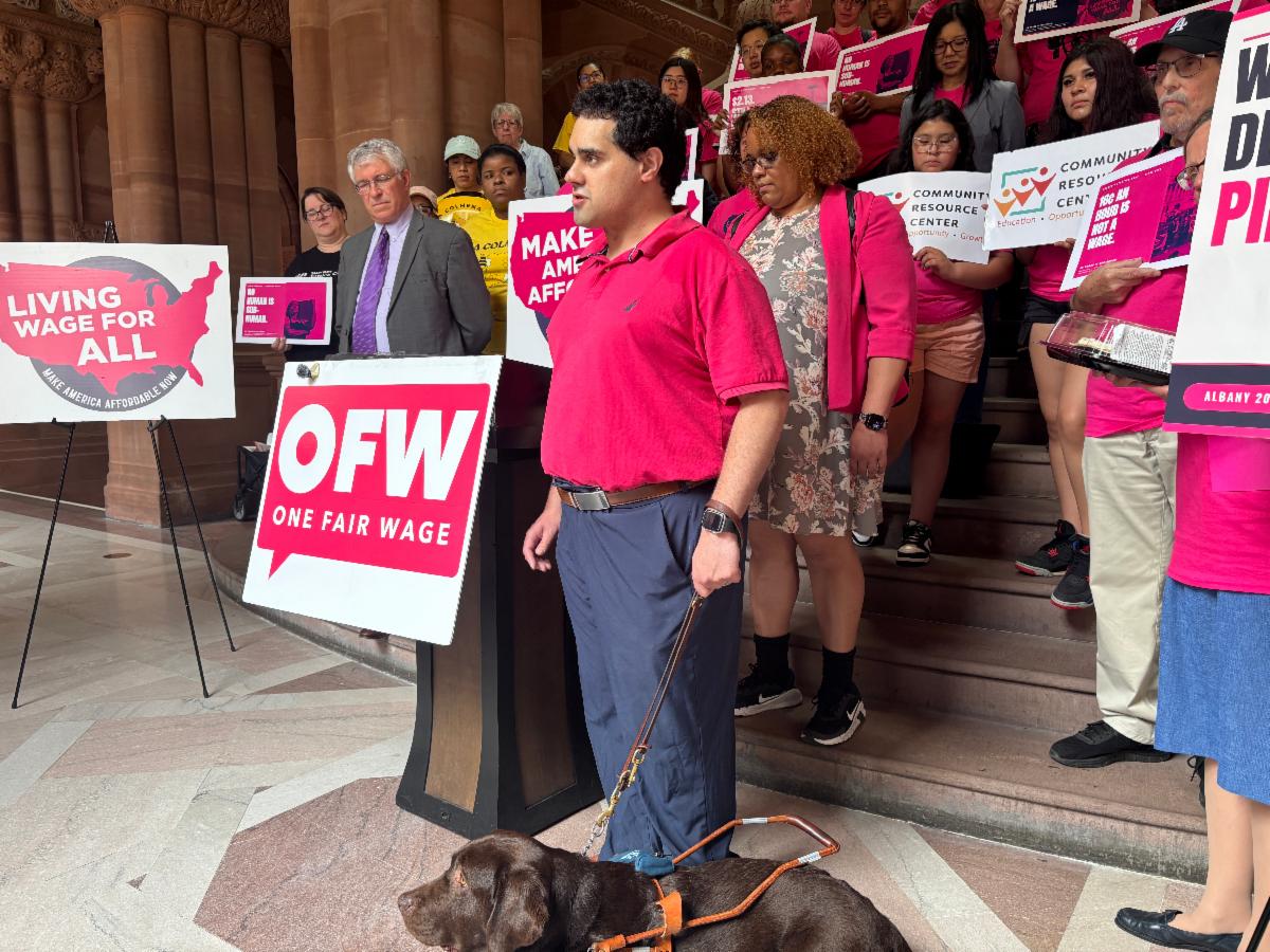 A blind man with his service dog, stands at a podium inside the New York State Capitol during a Living Wage for All rally, speaking in support of ending subminimum wages for disabled workers. His guide dog rests at his feet while advocates, disability rights supporters, and community members stand behind him holding signs promoting fair wages and economic justice.