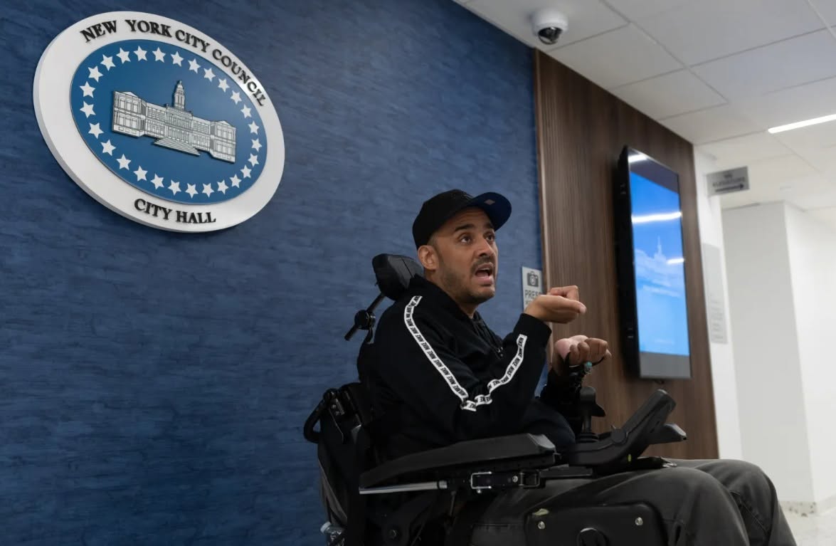 alt="José Hernandez sitting in his wheelchair in front of a blue wall with a sign of the New York City Council behind him."