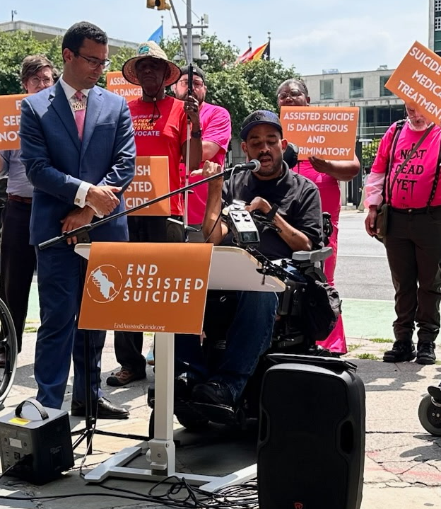 A wheelchair user speaks at an outdoor rally opposing assisted suicide as advocates hold protest signs behind him.
