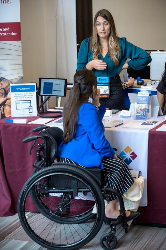 A woman in a wheelchair is at an exhibitor table speaking to a woman standing behind the table. 