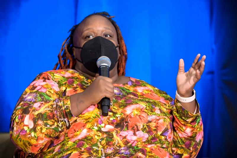 Keynote Speaker, Imani, sitting in a chair holding a microphone and speaking. She is wearing a mask. 