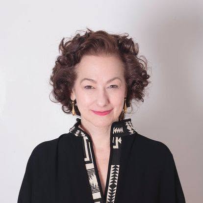 Portrait of Susan Dooha with short, curly brown hair wearing a black jacket with a patterned black-and-white collar, smiling against a light gray background.