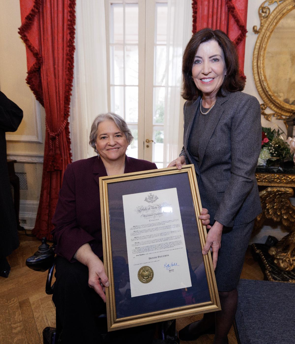 Executive Director, Denise Figueroa seated in a wheelchair accepting an award at the Women's History Luncheon from  Governor Hochul