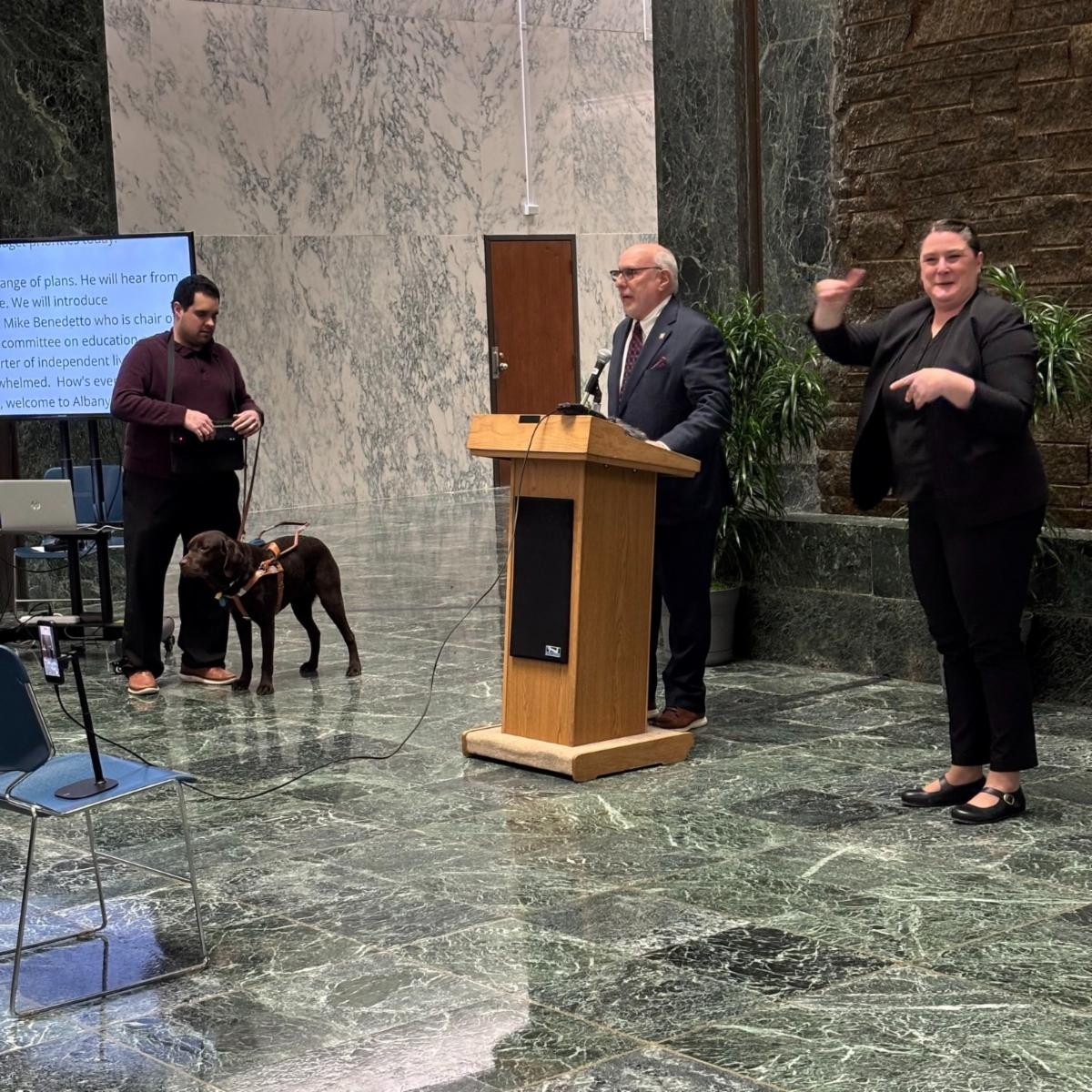 alt="Michael Benedetto speaks at a podium at the New York State Capitol, with a sign language interpreter and a service dog visible nearby during Budget Advocacy Day."