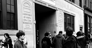 Black and white photo showing a group of people walking outside a building with classical architecture The building has large windows with engraved Latin text on stone panels reading A MONTIBUS AD MARIA UNA CUM SEDIMENTIS People appear to be standing in line or conversing near the entrance.