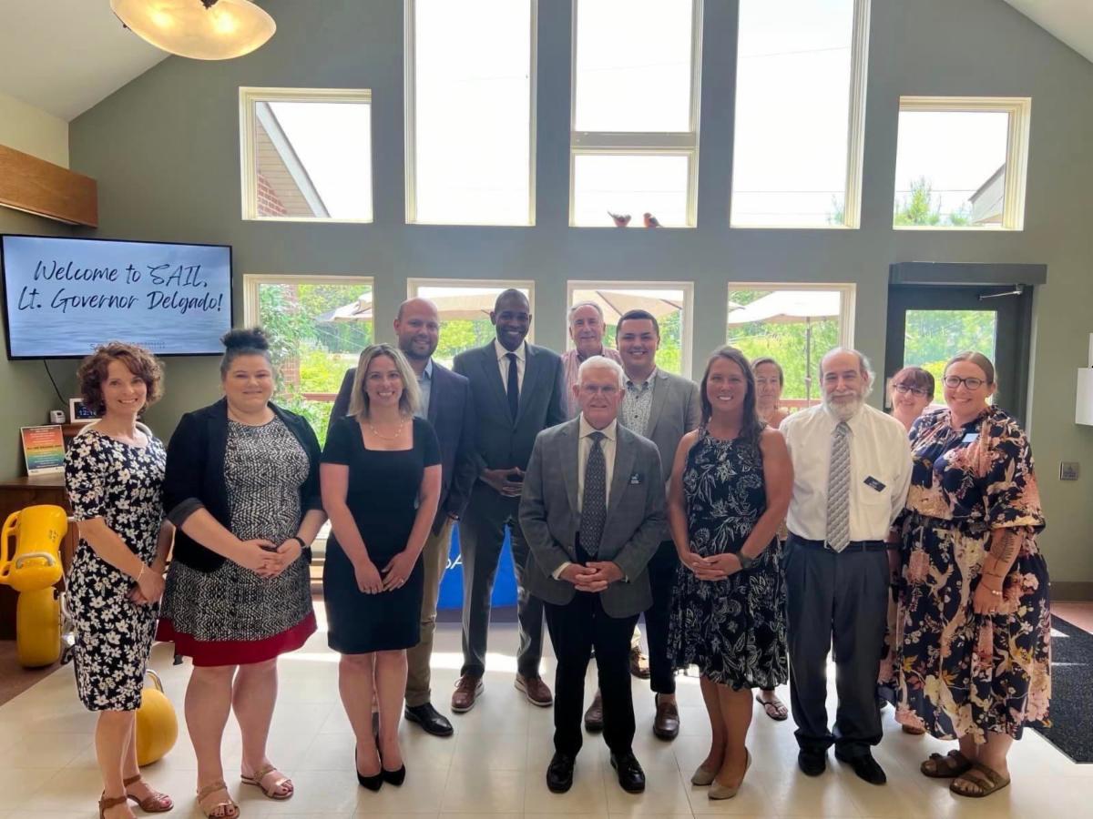 Group photo of Lieutenant Governor Antonio Delgado with Executive Director, Tyler Whitney, Director of Community Relations, Cam Cardinale, and staff smiling BIG and standing in front of a large set of windows that fill the entire wall, letting in a beautiful amount of natural light. To the left in the background, a TV screen reads, Welcome to SAIL Lt. Governor Delgado!