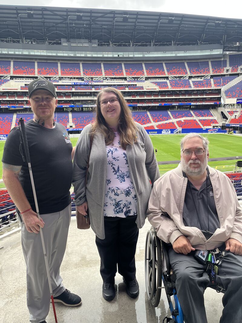 Aaron Baier, Stephanie Orlando and Todd Vaarwerk with WNYIL pose with the Buffalo Bills field and stadium, filled with red and blue chairs, in the backdrop. 