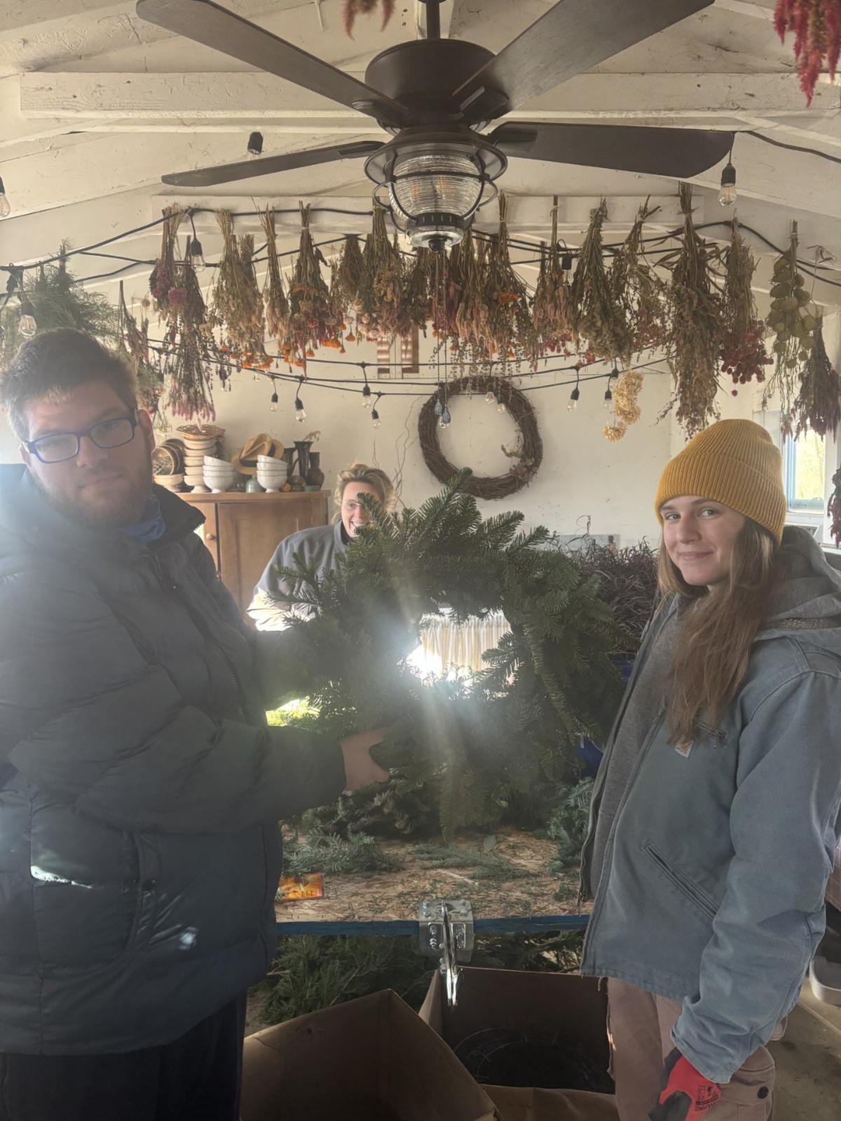Three people stand inside a cozy workshop or craft room. In the foreground, two individuals hold a wreath made of fresh pine branches. The person on the left wears a dark jacket and glasses, while the person on the right wears a yellow beanie, a light blue jacket, and red gloves. A third person stands in the background, partially obscured by the wreath, smiling and facing the camera. Overhead, a ceiling fan is surrounded by strings of dried herbs and flowers hanging from the ceiling. Shelves along the left wall hold neatly stacked bowls and jars. Natural light fills the room, creating a warm, creative, and welcoming atmosphere.