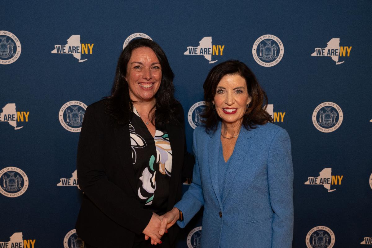 Lindsay Miller and Governor Kathy Hochul smiling and shaking hands in front of a backdrop with New York State logos and text.