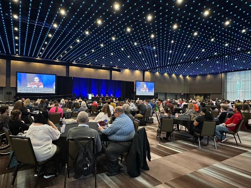 A conference room full of people sitting at tables watching and listening to a speaker. 