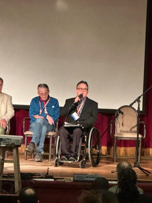 Doug sitting in a wheelchair speaking into a microphone while on stage.