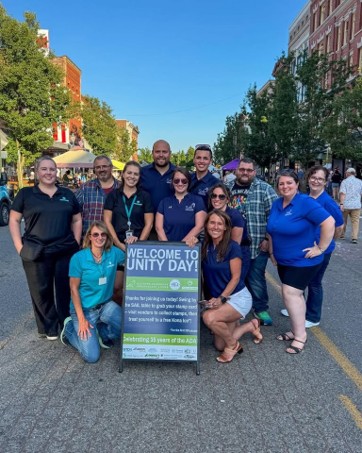 A group photo of eleven people smiling and posing on an open city street next to a vertical sign that reads "WELCOME TO UNITY DAY!" The individuals are dressed mostly in casual blue shirts and jeans. A woman in a turquoise shirt is kneeling on the left side of the sign, and another woman in a dark blue shirt is kneeling on the right side. The sign notes that the event is "Celebrating 35 years of the ADA" and invites attendees to collect stamps from vendors for a free Kona Ice. Buildings and other pedestrians are visible in the background under a clear blue sky