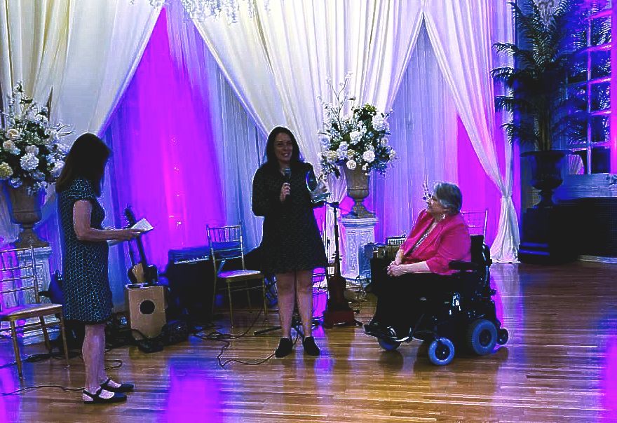Executive Director, Denise Figuero seated in a wheelchair giving an award to executive Director, Lindsay Miller at the 25th Dessert Spectacular
