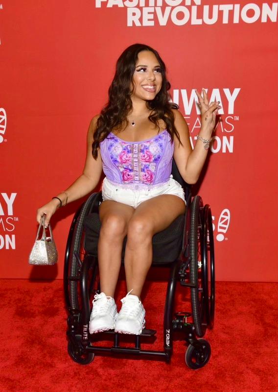 Mariah sitting in a wheelchair while smiling and holding up a peace sign. 