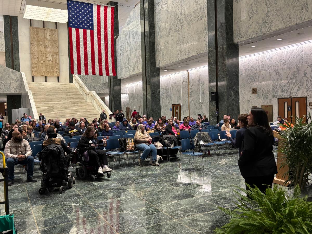 A large group of advocates seated in rows of blue chairs inside a grand marble atrium, likely at the New York State Capitol. An American flag hangs prominently from the ceiling above a wide staircase. In the foreground, a woman stands at a podium addressing the crowd. Several attendees use wheelchairs or mobility devices, and many wear purple shirts, suggesting coordinated advocacy efforts. The setting appears formal and civic, with tall marble columns and polished green marble floors.