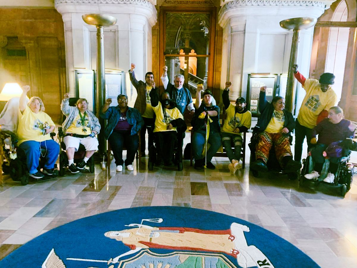 Group of advocates in the NYS Capital Building hands raised in the air, Nothing about us, Without us! 