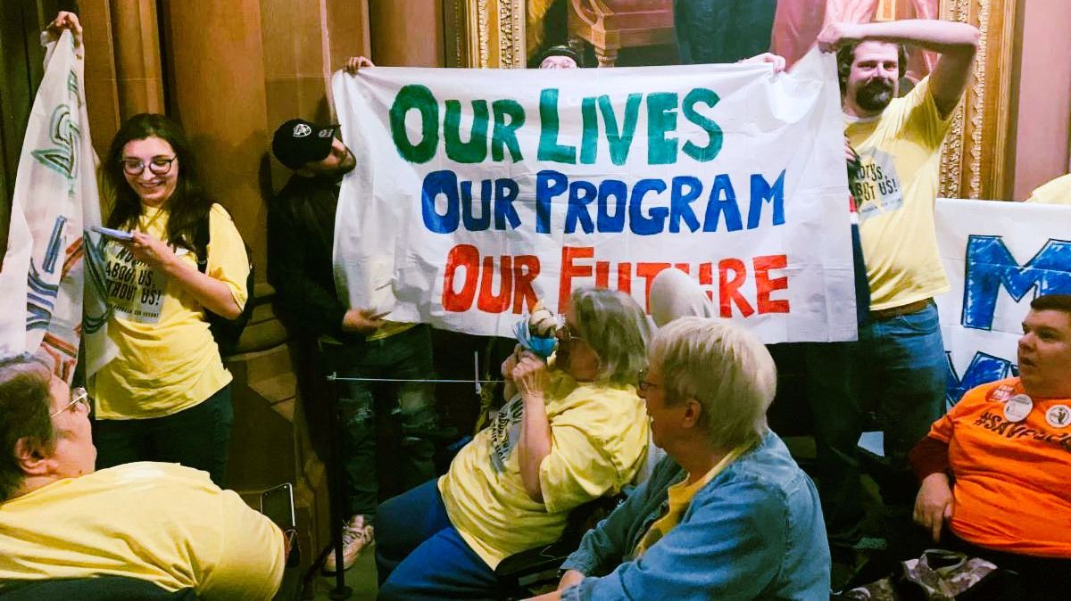 Group of 100 advocates in front of I Love NY sign. The photo shows people holding signs reading Fund Access to Home , CDPA is Essential, and Support Independent Living Funding while some chanted with fists in the air! 