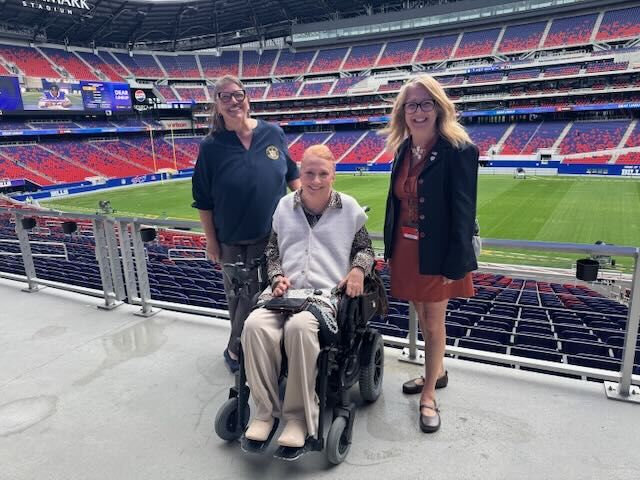 Holly Saupp, Kim Ridley and Kim Buchan with the office of the Chief Disability Office post at the Bills stadium with the field and scoreboard behind them. 