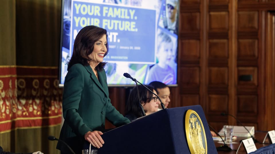 Governor Kathy Hochul speaks at a podium during a public event, addressing an audience while seated attendees listen behind her. A presentation screen in the background displays information about New York State initiatives.