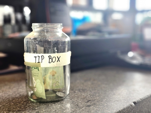 Close-up of a tip jar on a counter with some coins and a banknote inside_ labeled  Tip Box  in a caf  or restaurant setting.