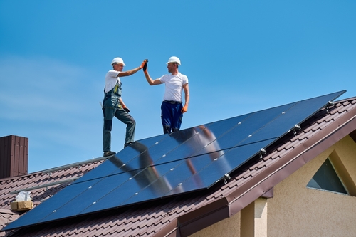 Worker building solar panel system on metal roof of house. Engineers giving high five to each other_ standing on rooftop while installing photovoltaic solar module outdoors.