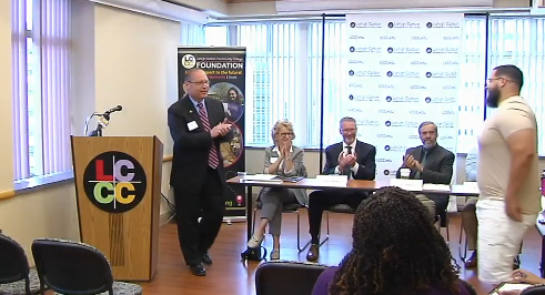 A speaker addresses an audience from a podium in a conference room at Lehigh Carbon Community College. A panel of three people sits at a table behind him under an LCCC Foundation banner.