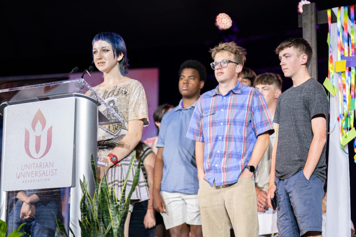 A youth speaker stands at a podium with the Unitarian Universalist Association logo, addressing an audience while several young people stand together behind them on stage during a General Assembly program.