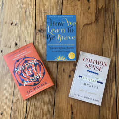 Three books are arranged on a rustic wooden floor. The book on the left is No One Left Alone by Liz Walker, with a coral-colored cover. At the top is How We Learn to Be Brave by Bishop Mariann Edgar Budde, featuring a blue cover with yellow and white text. On the right is Common Sense by Thomas Paine, with a white cover and blue lettering. The books are displayed in a loose triangle, suggesting recommended reading or featured titles.
